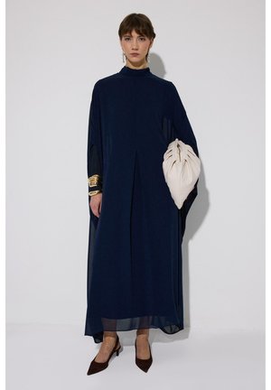 Nő hosszú sötétkék, hosszú ujjú ruhát visel, kezében egy nagy törtfehér neszesszer táskát tart, arany karkötőkkel és fülbevalókkal kiegészítve.