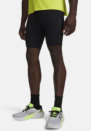 Atleet draagt zwarte strakke hardloopshorts, zwarte sokken en grijze neongele Under Armour hardloopschoenen op een witte achtergrond.
