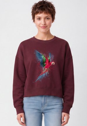 Jeune femme aux cheveux bruns courts porte un sweat-shirt bordeaux avec un motif coloré d'un perroquet volant, assorti à un jean bleu clair.