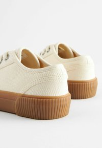 Płócienne sneakersy w kolorze kremowym z teksturowaną powierzchnią. Posiadają okrągłe oczka oraz podeszwę z gumy w kolorze karmelowym z pionowymi paskami.