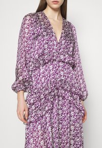 Robe violette avec imprimé floral blanc, décolleté en V profond, détails froncés et longues manches bouffantes. Tissu léger et transparent.