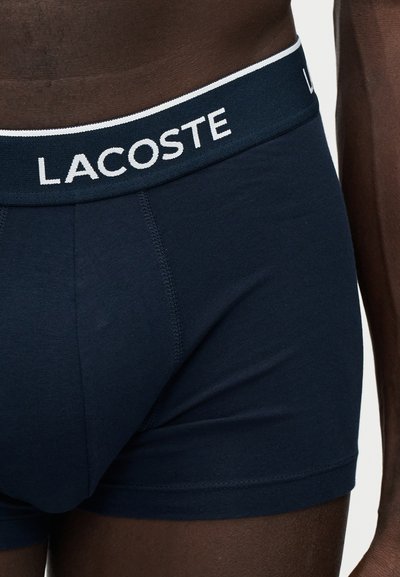 Крупен план на човек, носещ тъмносини боксерки на марката Lacoste с бяла лента на талията с лого.