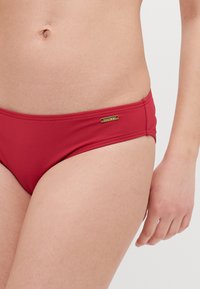 Rød bikiniunderdel laget av glatt, stretchy materiale, med et lavt liv og en liten gulllogo detaljer på siden.