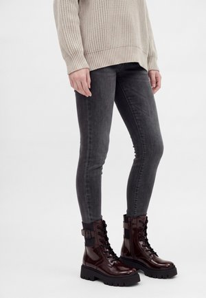Personne portant un pull en maille beige, un jean skinny gris foncé et des bottes lacées marron foncé brillantes avec des semelles épaisses et des boucles latérales.