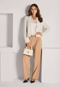 Frau in beigen Hose, Oberteil mit weißer Spitzenborte, cremefarbener strukturierter Jacke, mit Brille, die eine kleine cremefarbene Handtasche und schwarze High Heels in einem minimalistischen Umfeld hält.