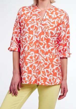 Žena v oranžovo-bílém květovaném topu s trojčtvrtečními rukávy vyhrnutými nahoru, v kombinaci s bledě žlutými kalhotami.