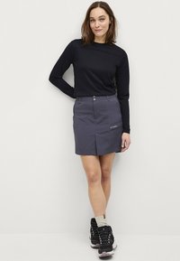 Grijze skort met een vlakke voorkant, twee knopen en een zijzak. Zwarte langemouwen shirt en zwarte wandelschoenen met grijze accenten.