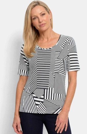 Blonde Frau trägt ein kurzärmeliges, schwarz-weiß gestreiftes Oberteil mit geometrischem Muster und steht vor einem einfachen hellen Hintergrund.