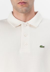 Homme portant un polo blanc côtelé avec trois boutons blancs et petit logo crocodile vert sur la poitrine gauche.