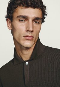 Polo a coste marrone con colletto a bottoni, caratterizzata da un dettaglio di bottone azzurro. Tessuto liscio e design aderente.