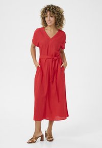 Röd linneklänning med V-ringning, korta ärmar och knytning i midjan. Har sidofickor och når till mitten av vaden. I kombination med bruna sandaletter med blockklack.