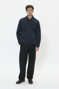 Chaqueta navy con dos bolsillos en el pecho, botones al frente y cuello puntiagudo; los pantalones negros de corte relajado y los zapatos marrones completan el conjunto.