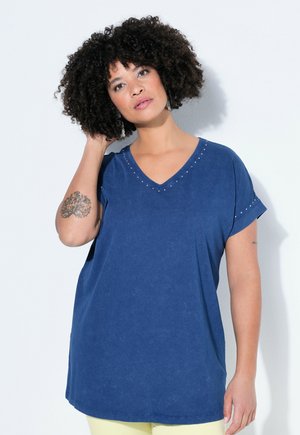 Kiharatukkainen nainen, jolla on sininen V-kauluksinen paita, jossa on pieniä niittejä, koskettaa hiuksiaan yhdellä kädellä valkoista taustaa vasten seisten.