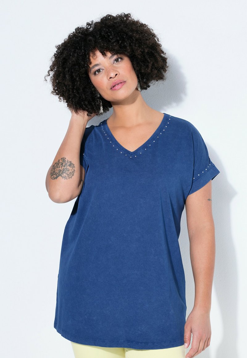 Frau mit lockigem Haar, die ein blaues V-Ausschnitt-Shirt mit kleinen Nieten trägt, mit einer Hand das Haar berührt und vor einem weißen Hintergrund steht.