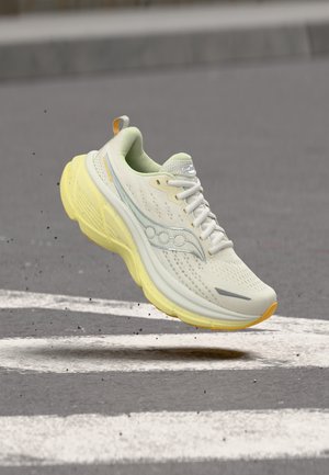 Világoszöld futócipő sárga talppal, amely az aszfalt felület felett lebeg, ahol fehér útjelzések láthatók.