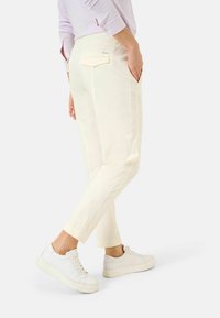 Osoba w jasnobeżowych, krótkich spodniach z tylną kieszenią i białych trampkach, stojąca z jedną nogą lekko uniesioną.