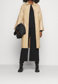 Imperméable beige brillant avec un col, des boutons-pression et des manches trois-quarts. Porté avec un haut noir, un pantalon large et des bottes noires.