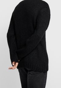 Svart stickad tröja med texturerad yta, långa ärmar och ribbat avslut. Buren över mörka jeans, sedd bakifrån.