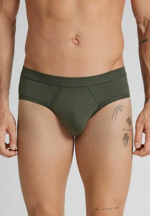 Mandlig overkrop iført olivengrønne briefs med synlige tatoveringer på øvre venstre lår og venstre underarm mod en neutral baggrund.