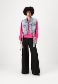 Gilet in denim sopra un felpa rosa brillante, abbinato a pantaloni neri a gamba larga. La modella tiene una piccola borsa rosa e indossa scarpe nere.