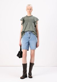 Grøn blouse med polkaprikker og volangærmer, lyseblå denimshorts og sort blomsterpose. Knæhøje brune læderstøvler med spænder.