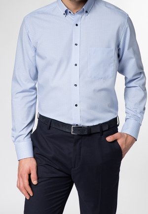 Uomo che indossa una camicia da uomo a quadri azzurri infilata in pantaloni neri con cintura nera, con una mano in tasca, davanti a uno sfondo neutro.