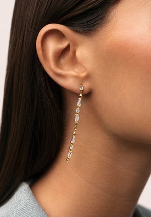 Oreja derecha de mujer con un pendiente de gota dorado con gemas rectangulares transparentes, cabello liso castaño oscuro y tono de piel claro visible.