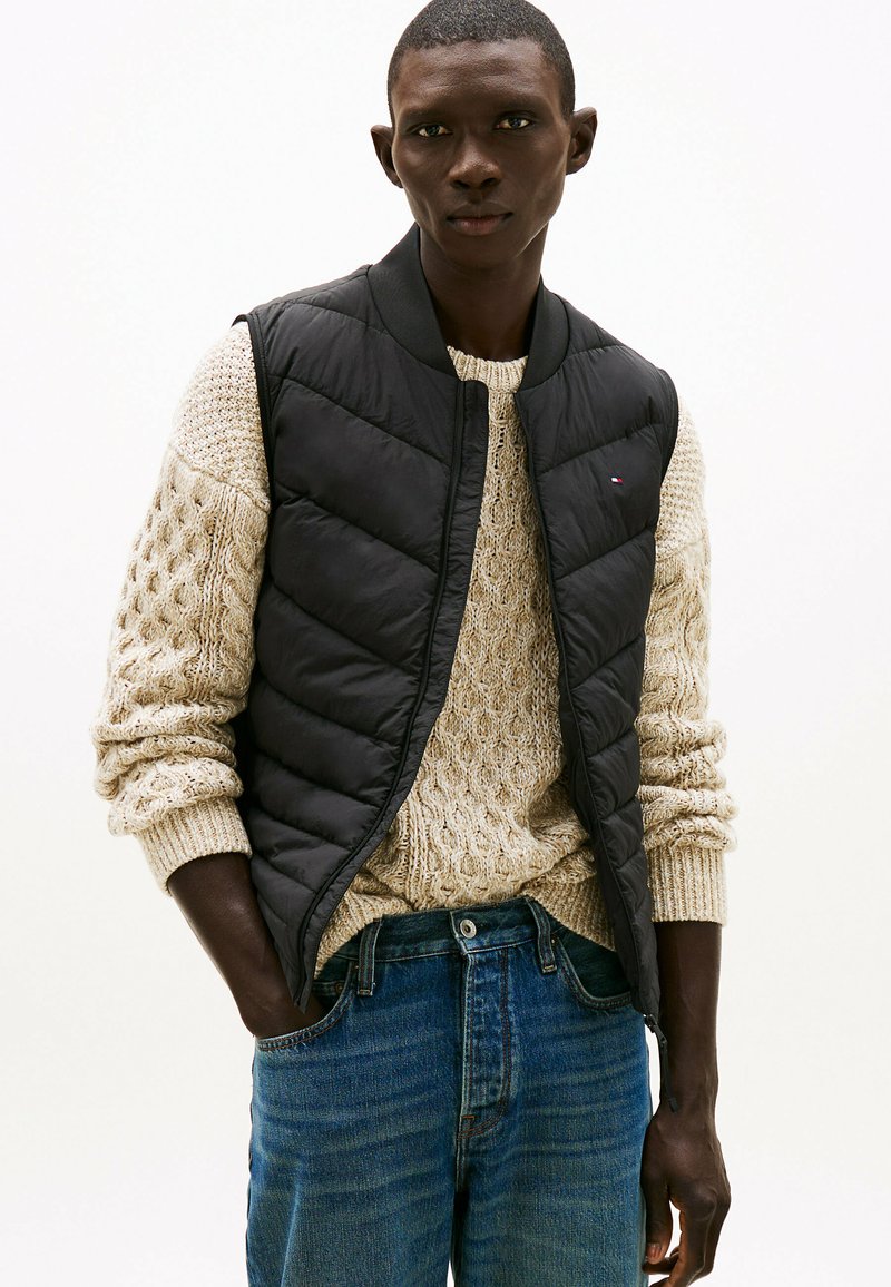 Chaleco negro acolchado con un cuello redondo, llevado sobre un suéter grueso de punto trenzado en color beige, combinado con jeans de pierna recta azules.