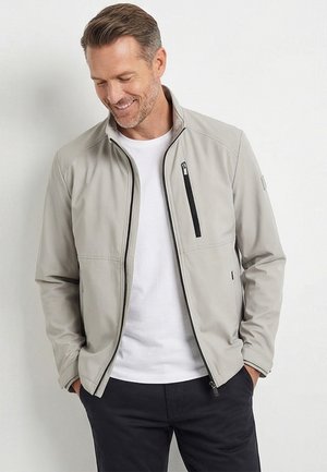 Lachende man die een lichtgrijze jas met rits draagt over een wit shirt, met zijn handen in de zakken, tegen een effen witte achtergrond.