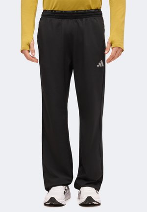 Pantalon de sport noir à coupe large avec une taille élastique et un logo blanc sur la cuisse, porté avec des baskets blanches et un haut à manches longues jaune.
