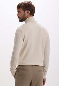 Mann mit braunen Haaren und Bart, der einen cremefarbigen strukturierten Rollkragenpullover und beige Hosen trägt, nach links blickend mit den Händen in den Taschen.