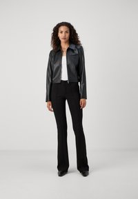 Schwarze Kunstlederjacke mit einem kragenlosen Halsauschnitt, vorderem Reißverschluss und einer einzelnen Brusttasche, kombiniert mit schmalen schwarzen Hosen.