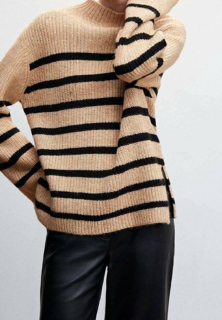 Pull en tricot beige avec des rayures horizontales noires, porté avec un pantalon noir, un bras plié et la main près du visage.