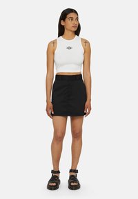 Witte cropped tanktop met een logo, gecombineerd met een zwarte minirok met een riem en een knoopsluiting aan de voorkant. Zwarte sandaaltjes met bandjes.