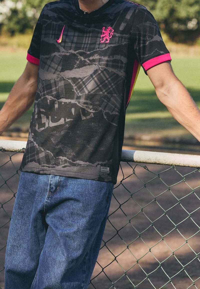 T-shirt nera a fantasia con accenti rosa, caratterizzata da un logo di leone e un design a collo rotondo. Abbinata a jeans blu, appoggiata a una recinzione.