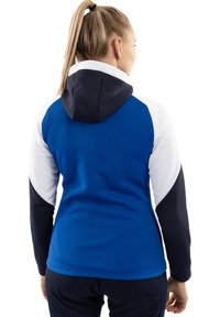 Giacca con cappuccio caratterizzata da pannelli blu, bianchi e blu scuro. Tessuto morbido con maniche lunghe e design aderente. La vista posteriore mette in risalto il contrasto dei colori.