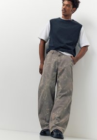 Fiatal férfi fekete ujjatlan pulóvert visel fehér póló fölött, szürke, absztrakt mintás bő szárú nadrágot és fekete cipőt, áll.