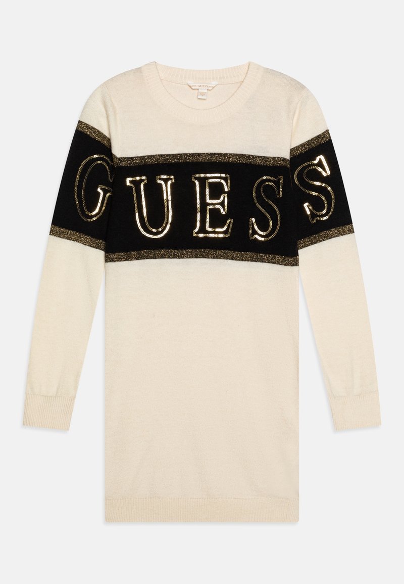 Longue robe pull crème avec une bande horizontale noire sur la poitrine et le haut des manches, ornée d’un grand lettrage doré à sequins « GUESS ».