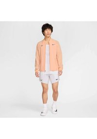 Chaqueta de cremallera color melocotón claro con una textura suave, combinada con una camiseta sin mangas blanca y shorts blancos, que presentan logotipos y acentos negros de Nike.