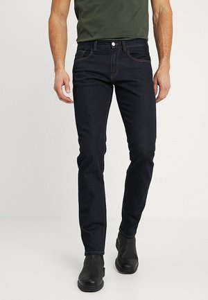 Mann, der schlanke dunkelblaue Jeans, schwarze Knöchelstiefel und ein olivgrünes T-Shirt trägt, steht vor einem schlichten hellen Hintergrund.