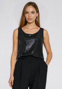 Młoda kobieta z długimi włosami, ubrana w czarny, błyszczący top bez rękawów i czarne spodnie z wysokim stanem, stoi na jednolitym tle.