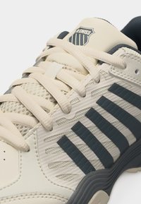 Μπεζ αθλητικό παπούτσι K-Swiss με μαύρες ρίγες, μπεζ κορδόνια, λεπτομέρειες από δίχτυ και το λογότυπο K-Swiss στη γλώσσα.