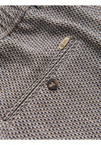 Gros plan d'un tissu pied-de-poule marron et crème avec un bouton marron foncé et une petite étiquette en laiton portant la mention "Ombre Heritage."