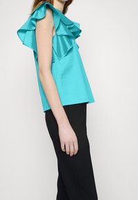Top turquesa con mangas con volantes, textura suave, corte holgado, combinado con pantalones negros, con un brillo sutil en los volantes.