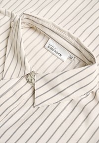 Blusa crema con sottili righe nere, dotata di un colletto classico e un bottone tondo texturizzato. Etichetta visibile: "Karen by Simonsen."