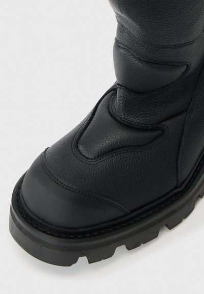 Schwarze Lederstiefel mit strukturierten Oberflächen, abgerundeter Zehenpartie, gepolsterten Details und kontrastierender, dicker Gummisohle. Branding auf der Seite sichtbar.