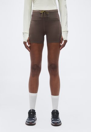 Personne portant un short marron taille haute avec un cordon, un haut blanc à manches longues avec des trous pour les pouces, des chaussettes blanches montantes et des baskets noires.