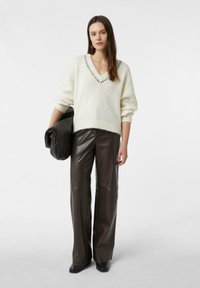 Kremowy sweter z dekoltem w serek z prążkowanym wykończeniem, oversize'owe rękawy, w połączeniu z wysokim stanem szerokimi spodniami z brązowej sztucznej skóry. Modelka trzyma teksturowaną torbę.