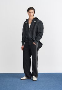 Jacket com padrão xadrez preto e fecho éclair, com capuz, combinado com calças largas pretas com uma faixa lateral branca e ténis brancos.