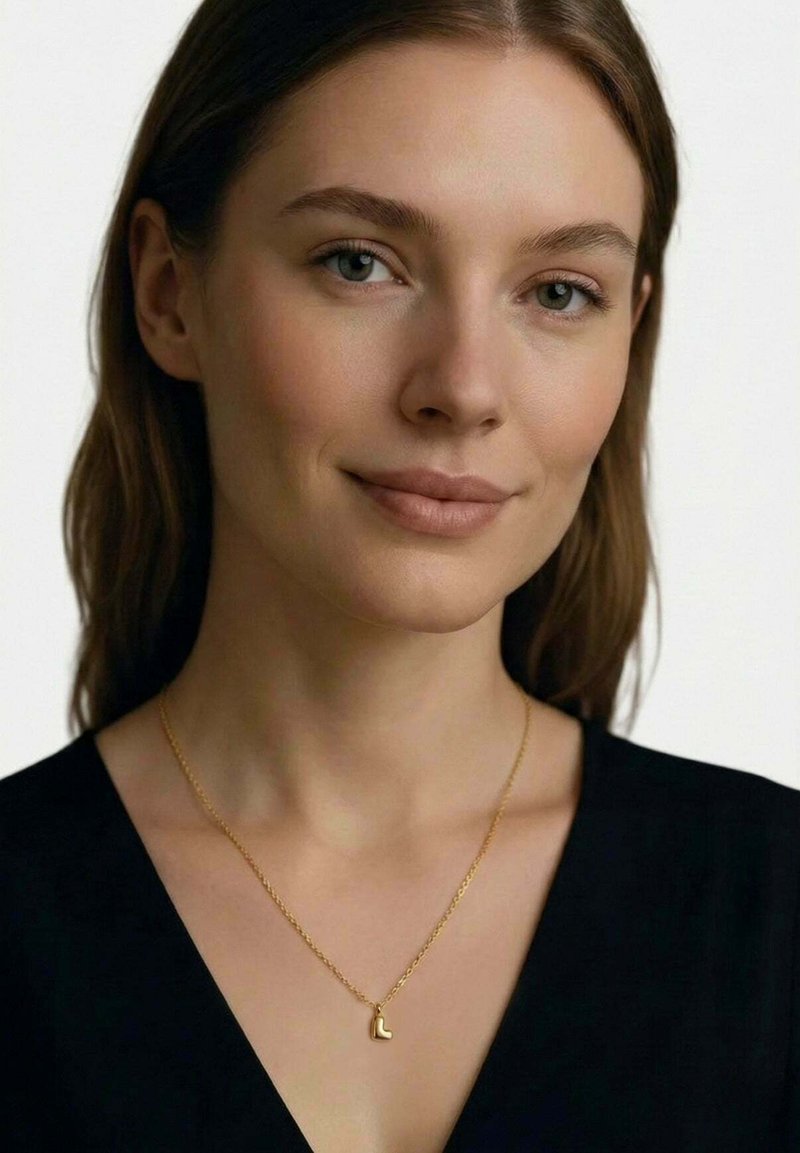 Junge Frau mit heller Haut und braunen Haaren trägt ein schwarzes Oberteil und eine goldene Halskette mit einem kleinen Anhänger in Form eines "L".